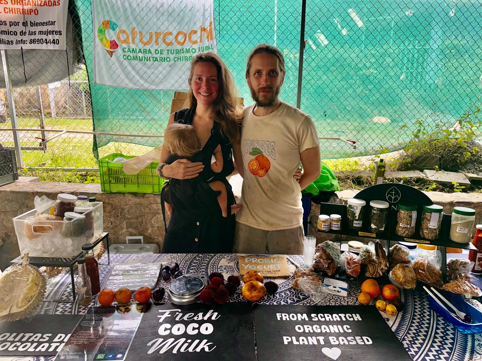 Manna's founders Jonas and Lydia with their son at a Southern Costa Rica feria, behind a stall of raw vegan foods, fresh fruit and ferments